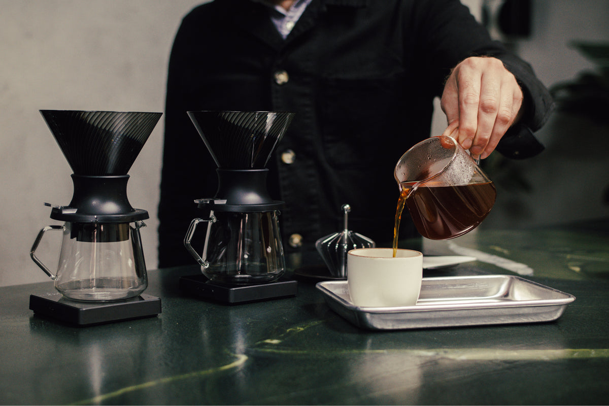A person in a black jacket pours freshly brewed coffee from a glass carafe into a white cup on a metal tray, with two black pour-over coffee makers on a dark green marble countertop in the background.