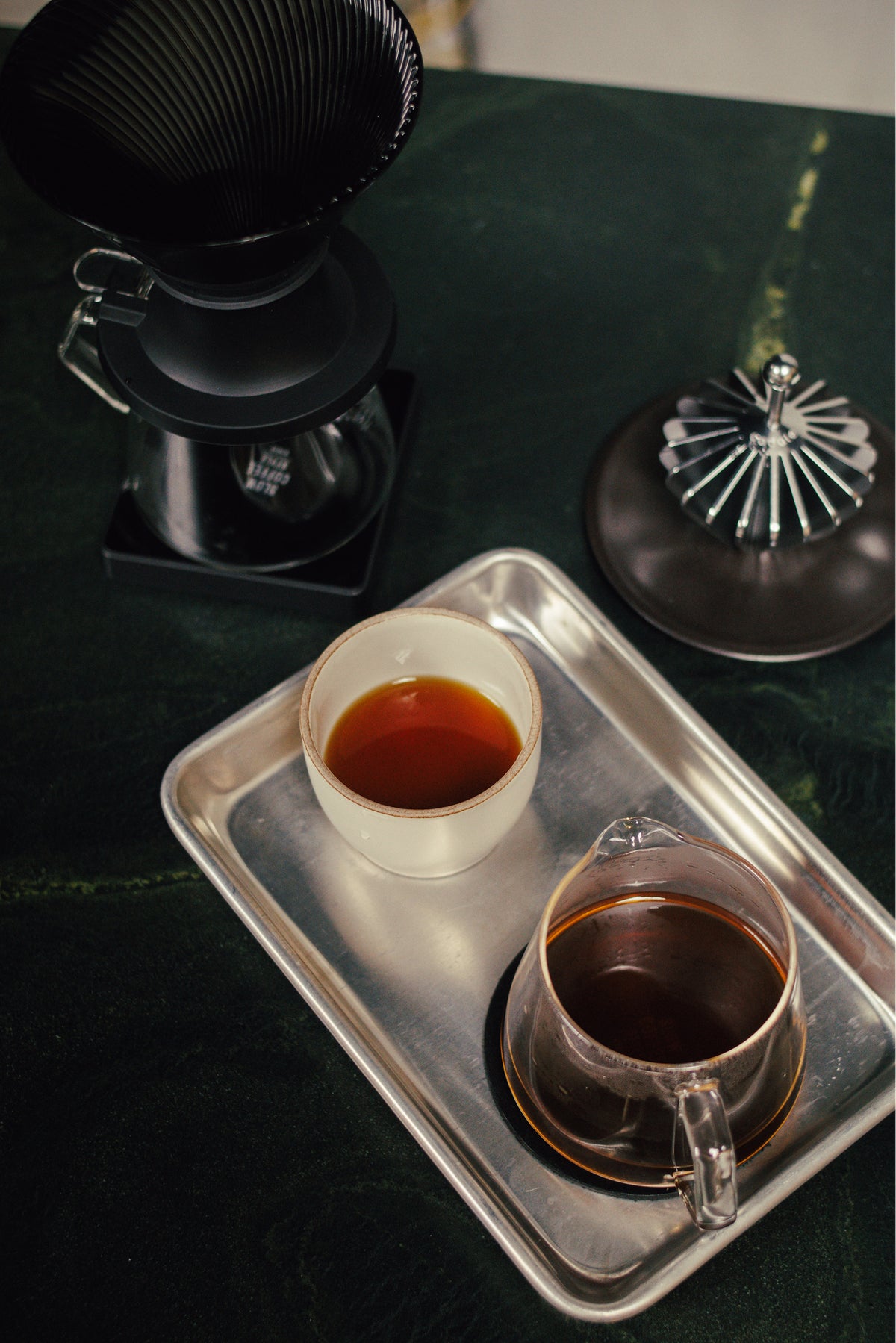 A coffee brewing setup on a dark green table, featuring a black pour-over dripper, a glass carafe filled with dark coffee, a white cup of lighter coffee, and a metal tray.